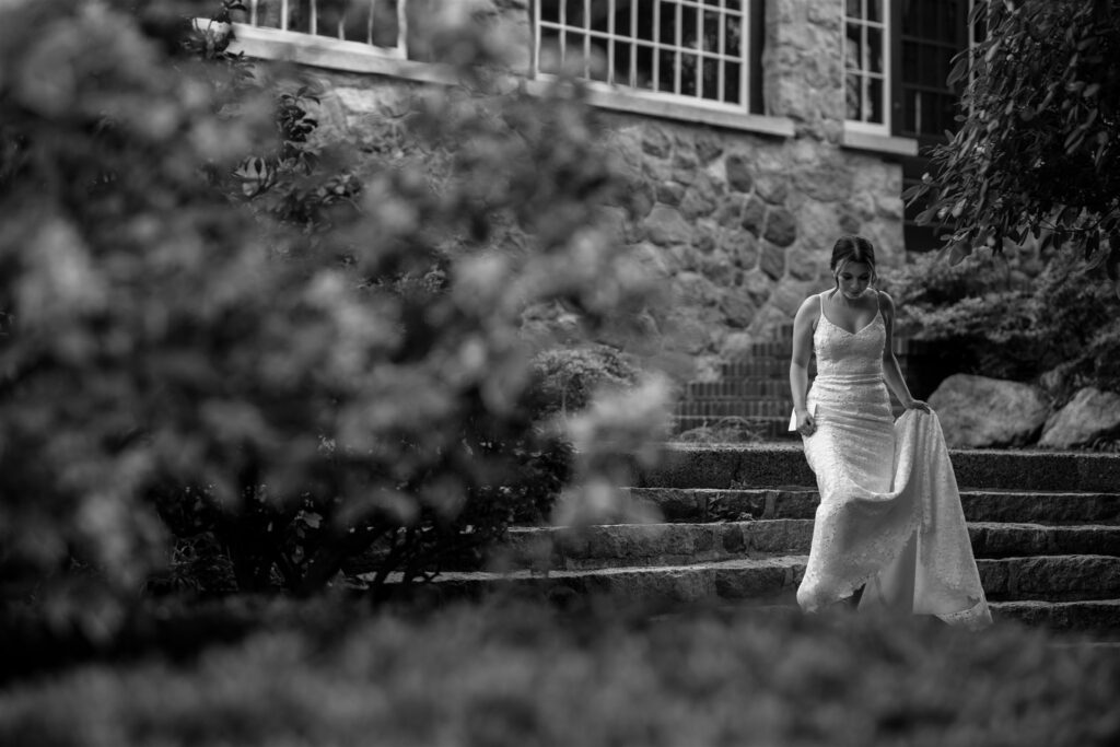 A black and white photo showing a bride in a white lace dress walking down stone stairs at her wedding. 
